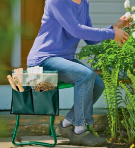  This image shows an elderly woman wearing a lilac long-sleeved top and pale jeans sitting on a garden kneeler. She is pruning a bush with white flowers. She has an array of gardening tools in the side pockets of the kneeler which is dark green in colour