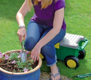 Tools for elderly gardeners. This image shows a gardening trolley. It is a wheeled trolley with a seat. The wheels are black plastic with a yellow centre circle. The trolley is green with a pale grey lid. It shows a woman sat tending a large blue flower pot.