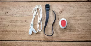 This image shows a call bell system pendant It is a small oval device which is white. It has a clear red button in the centre. Also shown is a black wrist strap and a white lanyard. 