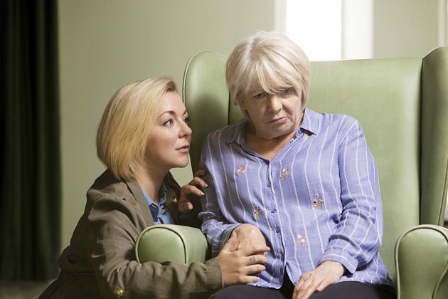 this image shows a woman sat in a large green chair who has had a stroke. Her Daughter kneels beside her