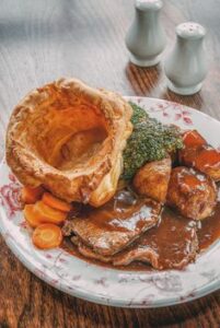 Good diet for elderly people. This image shows a large yorkshire pudding, sliced roast beef, broccoli, sliced carrots and gravy.