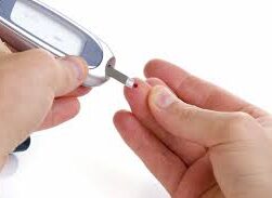 this image shows a person performing a diabetic prick test to test blood sugar levels. It shows a glucose testing machine and a testing strip it also shows a drop of blood on the finger next to the persons ring finger.