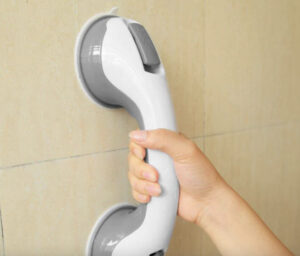Suction grab bars. This image shows a grey and white coloured suction grab bar. It is stuck onto beige tiles and someone is grabbing hold of it with their right hand