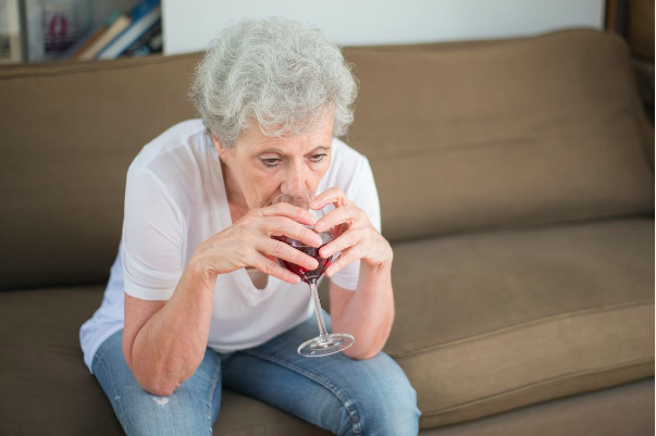 Alcohol addiction among older adults. This image shows an older lady with grey hair holding a glass of red wine with both hands. She is sat on a brown sofa and is wearing a pink top and pale blue jeans