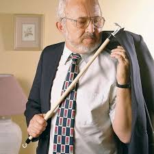 Dressing yourself with limited mobility is easier with a dressing stick.This image shows an older man putting on a dark suit jacket using a dressing stick. The stick is about a meter long with a hook on one end. The man is wearing a white shirt and a patterned darker tie. He has grey hair and beard and is wearing glasses. He is in his bedroom
