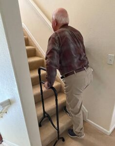 Walking stick for stairs. This image shows an elderly gentleman with a walking stick that fits onto two stair treads. It is rectangular in shape and has a wide handle for stability. The gentleman is using it on a staircase. He is wearing a check shirt and light coloured trousers. He has grey hair and is balding. The walls of the staircase are white and the stair carpet is caramel.