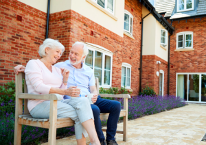Choosing the perfect retirement home. This image shows an elderly couple sat outside on a wooden bench outside a red brick building. The couple are chatting and looking happy. It is Summer and it is bright and they are dressed in lightweight clothes 