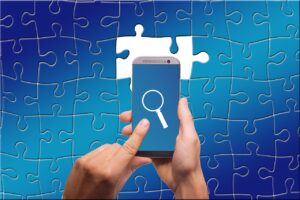 Dementia Diagnosis and Treatment . Image shows a blue jigsaw puzzle with two missing pieces in the centre. These are white. Above the puzzle is a persons hands holding a mobile phone. The phone screen is also blue and there is a white magnifying glass shown. Searching for the missing pieces. 