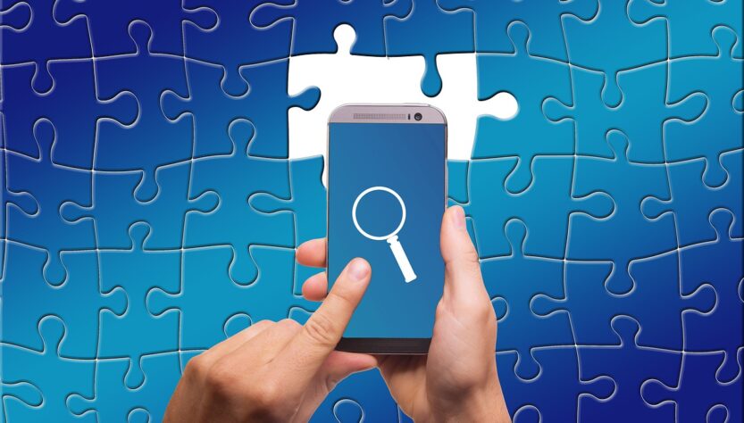 Dementia Diagnosis. Image shows a blue jigsaw puzzle with two missing pieces in the centre. These are white. Above the puzzle is a persons hands holding a mobile phone. The phone screen is also blue and there is a white magnifying glass shown. Searching for the missing pieces.