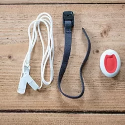 This image shows a Waterproof call bell pendant. It is a small oval device which is white. It has a clear red button in the centre. Also shown is a black wrist strap and a white lanyard.