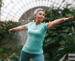 Maintaining mobility in Old Age. This image shows an older woman in a light green, short-sleeved tee shit. She is wearing blue leggings and has her arms outstretched to the front and back. She has short grey hair and looks happy