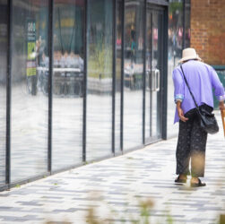 Article Title: Muscle damage recovery. This image shows an elderly woman walking along a patterned pavement. She is using a stick and is slightly hunched. She is wearing a purple jacket, blue jeans, sandals and a hat. She has a dark handbag slung across her back. To he side of her are tall rectangular windows that look like mirrors.