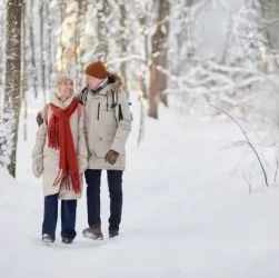 Financial help this Winter. This image shows an elderly couple walking in a wood. There is heavy snow on the ground and on the trees. They are wrapped up against the cold and the woman is wearing a bright red knitted scarf. They both have hats on.