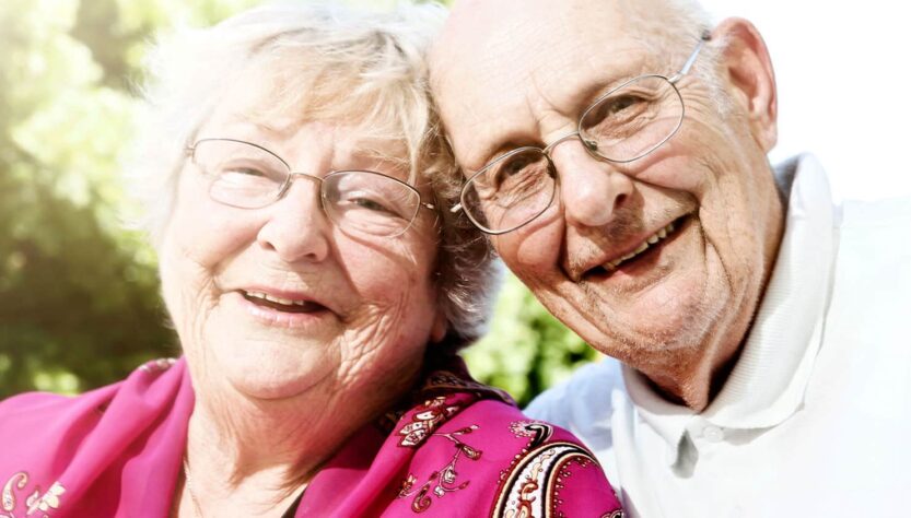 Caring for a parent. This image shows an older man and woman. They both have white hair and are smiling and looking happy. The woman is wearing a bright fushia pink cardigan and the man a white polo shirt