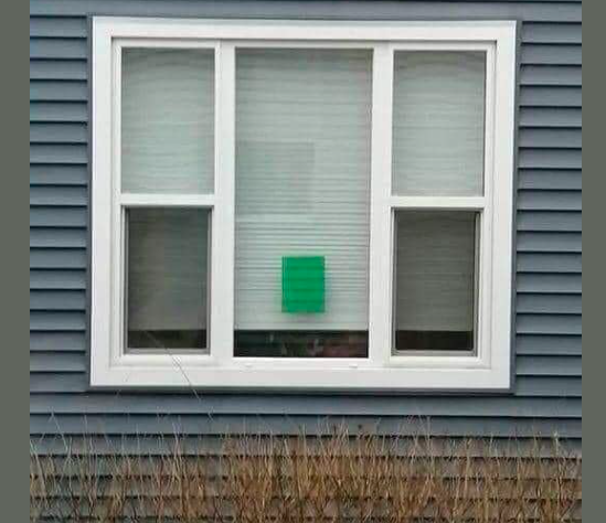 This image is shows a large white window pane. It has one large single centre panel and two equal length ones divided in two. In total there are 5 windows. Their is a window blind and it is almost to the window sill. Just above the window sill is a green sheet of card attached to the window. The card is part of a simple alert system to allow elderly people to signal whether they are OK or need help