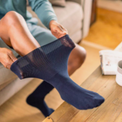 This image shows a woman demonstrating a pair of socks specially designed for people with Lymphoedema. The socks are navy in colour and she is clearly demonstrating the ability of the socks to stretch far more widely at the top than standard socks.