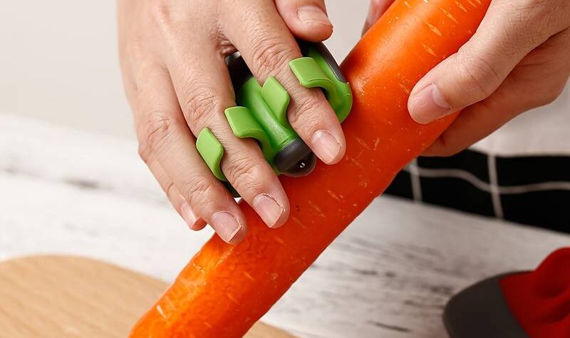 Peeler for arthritic hands. This image shows a peeler you slide your fingers into to peel veg. The one shown has green finger guides. The person is holding a large carrot and it is resting on a wooden chopping board.